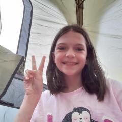 A girl with shoulder-length brown hair is smiling and holding up a peace sign. She's wearing a pink t-shirt with a cartoon penguin on it and is in a tent.