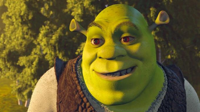 A close-up of Shrek smiling with a slight smirk. He's green with red eyes, wearing a brown vest. The background shows blurry green trees and bright light.