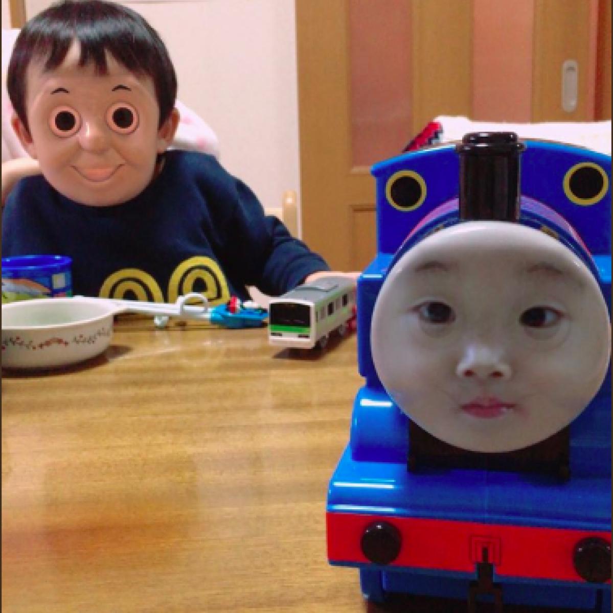 A child with a human face on his head and a toy train with a human face on the front of it are on a table together.