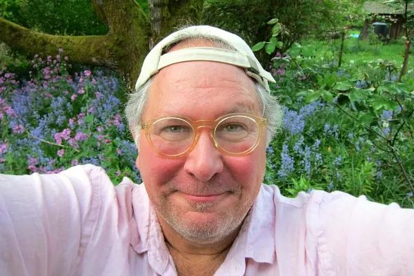 A man with glasses and a white cap is taking a selfie in front of some flowers.