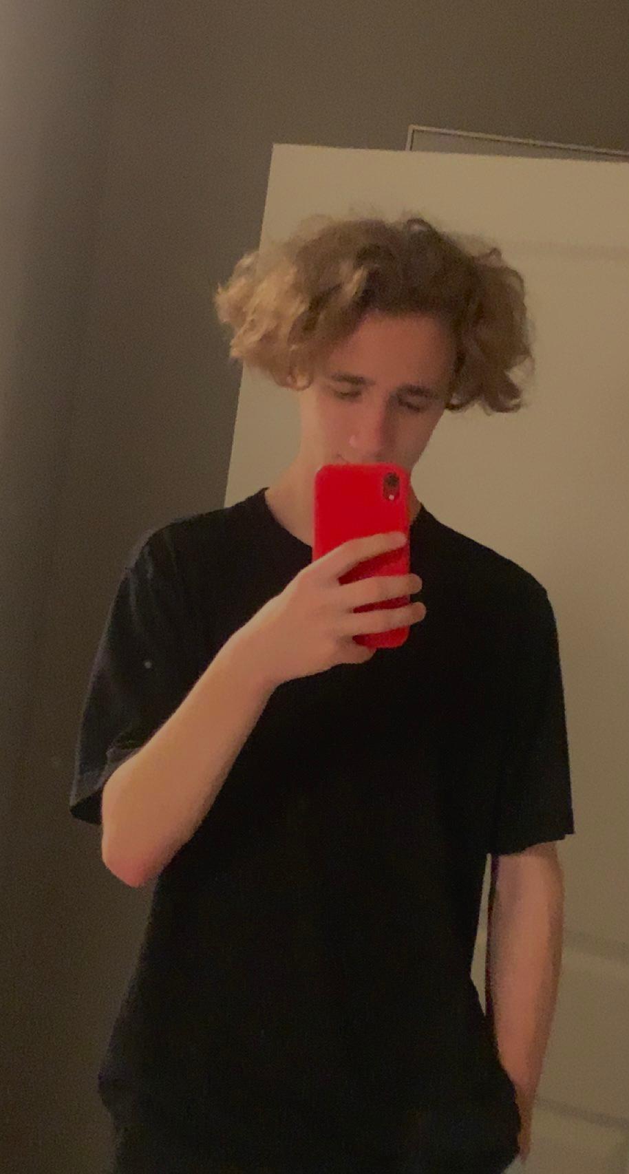 A young man with curly blond hair is taking a selfie in the mirror, holding a red phone.