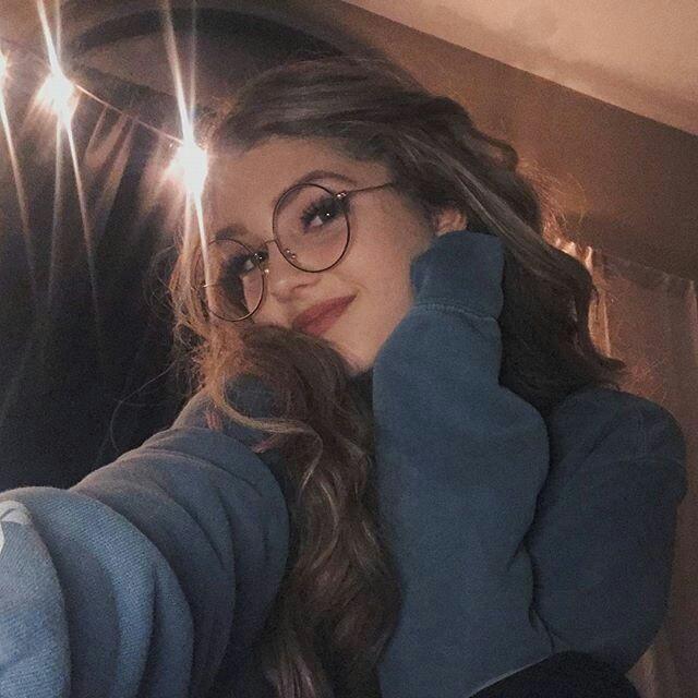 A girl with wavy brown hair wearing glasses and a blue sweatshirt looks at the camera. The image has a slightly soft focus and uses warm lighting.