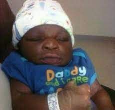 A close-up photo of a baby with dark skin wearing a white cap and a blue t-shirt that says "Daddy Love."