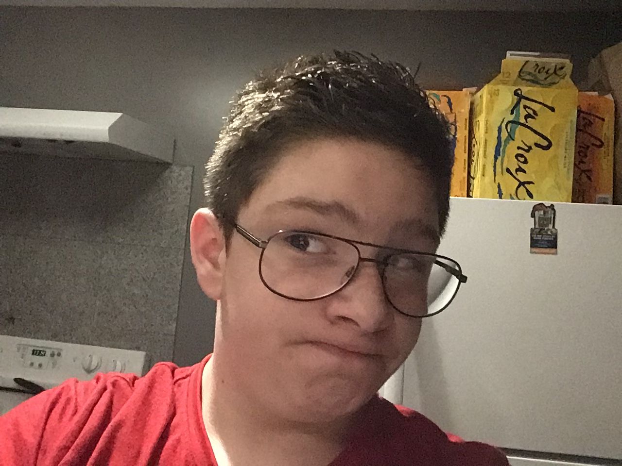 A photo of a young person with short, dark hair and glasses, making a slightly confused face. There are several boxes of La Croix sparkling water on the shelf behind them.