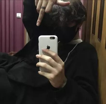 A person with dark curly hair and a black mask is holding a white phone up to take a selfie. They have a black long-sleeved shirt on and a ring on their right hand. They are also wearing a necklace.