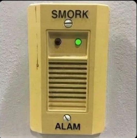 A yellow rectangular alarm device with the text "SMORK" on top and "ALAM" at the bottom. Two small light bulbs are in the center, the right one green.