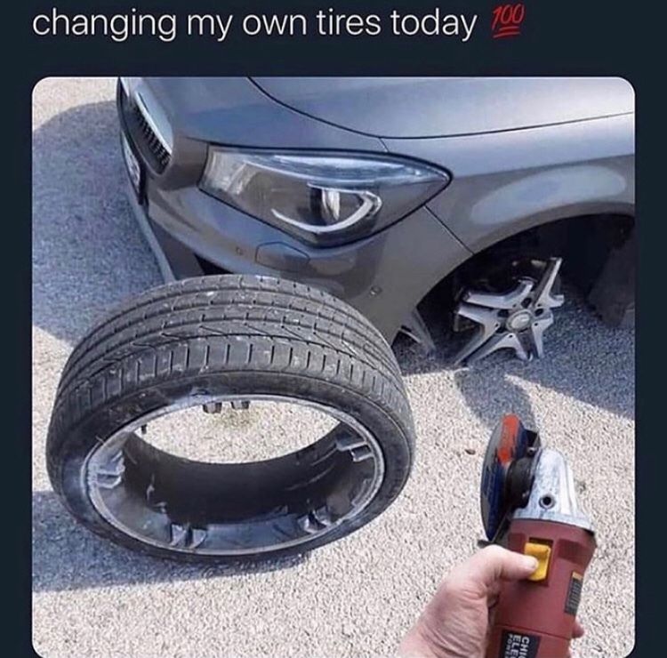 A car is missing one tire. A tire is laying on the ground in front of the car. A hand is holding a power tool, likely a grinder, in front of the tire.