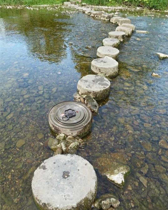 A picture shows a small river or creek with stepping stones. The stones are placed in a line across the water. Most stones look like concrete cylinders. One stone is a metal lid or pot cover.