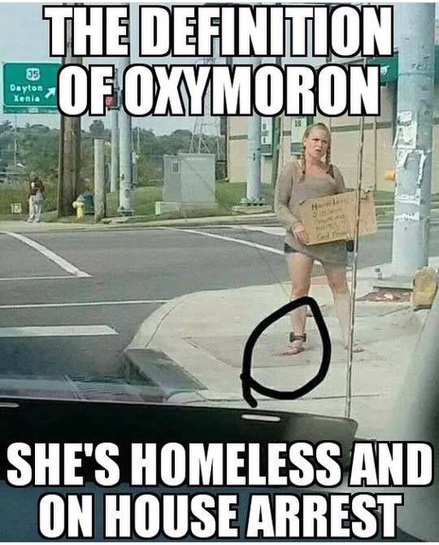 A woman is standing at the side of the road holding a cardboard sign. She is wearing an ankle monitor. The text on the image says: “The definition of oxymoron. She's homeless and on house arrest.”