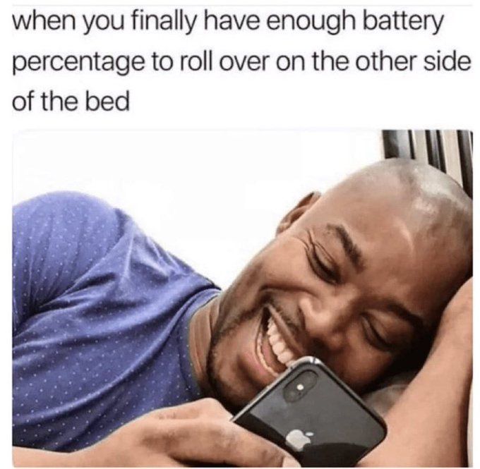 A man is smiling while looking at his phone in bed. The text says, "when you finally have enough battery percentage to roll over on the other side of the bed."