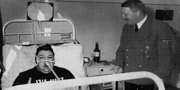 A black and white photo shows a person with a feeding tube lying in a hospital bed. Standing near the bed is Adolf Hitler, wearing his uniform. There is a bottle of wine on the nightstand.