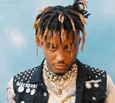 A portrait of Juice WRLD with a serious expression, wearing a studded vest and multiple necklaces, against a light blue background.