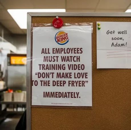 A cork board with two papers pinned to it. The left paper has the Burger King logo at the top. Below is the text: "ALL EMPLOYEES MUST WATCH TRAINING VIDEO 'DON'T MAKE LOVE TO THE DEEP FRYER' IMMEDIATELY." To the right is a small get well note that says "Get well soon, Adam!"