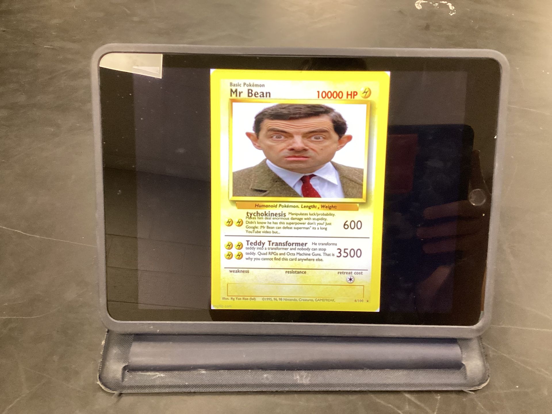 A photo of a tablet on a table displaying a custom-made Pokemon card of Mr. Bean. The card lists his name, HP, and two attack abilities with descriptions and attack points.