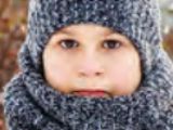 A close-up of a child's face, wearing a gray knitted hat and scarf. The child's eyes are looking directly at the viewer, and their nose and mouth are mostly covered by the scarf. The background is blurry, suggesting an outdoor setting.