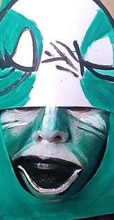 A person wearing green face paint and a white headpiece with black markings, resembling a mask.