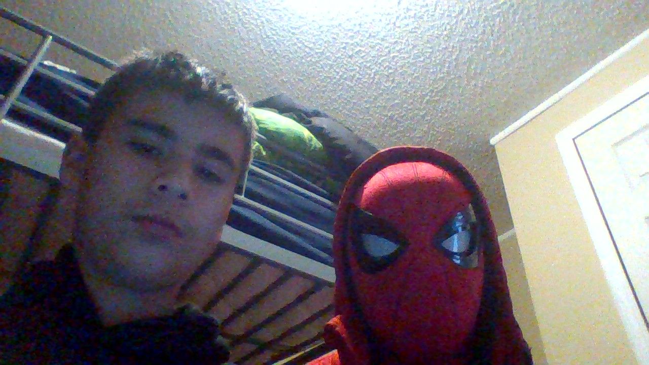 A photo shows two people. One on the left is a boy. He looks at the camera, he has short brown hair and a serious face. On the right is someone wearing a Spiderman costume with the mask on. They are in an indoor setting with a bunk bed in the background.