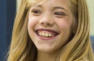 A close-up of a young blonde girl with a wide smile, showing her teeth. She looks happy and has rosy cheeks.