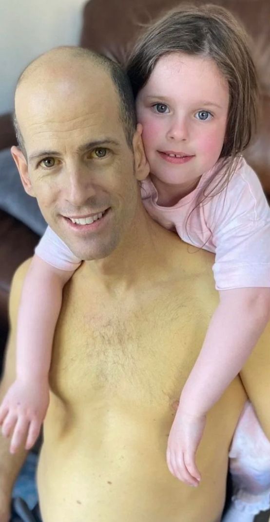 A father and daughter are posing for a picture. The father is shirtless, and the daughter is behind him, her arms around his shoulders.