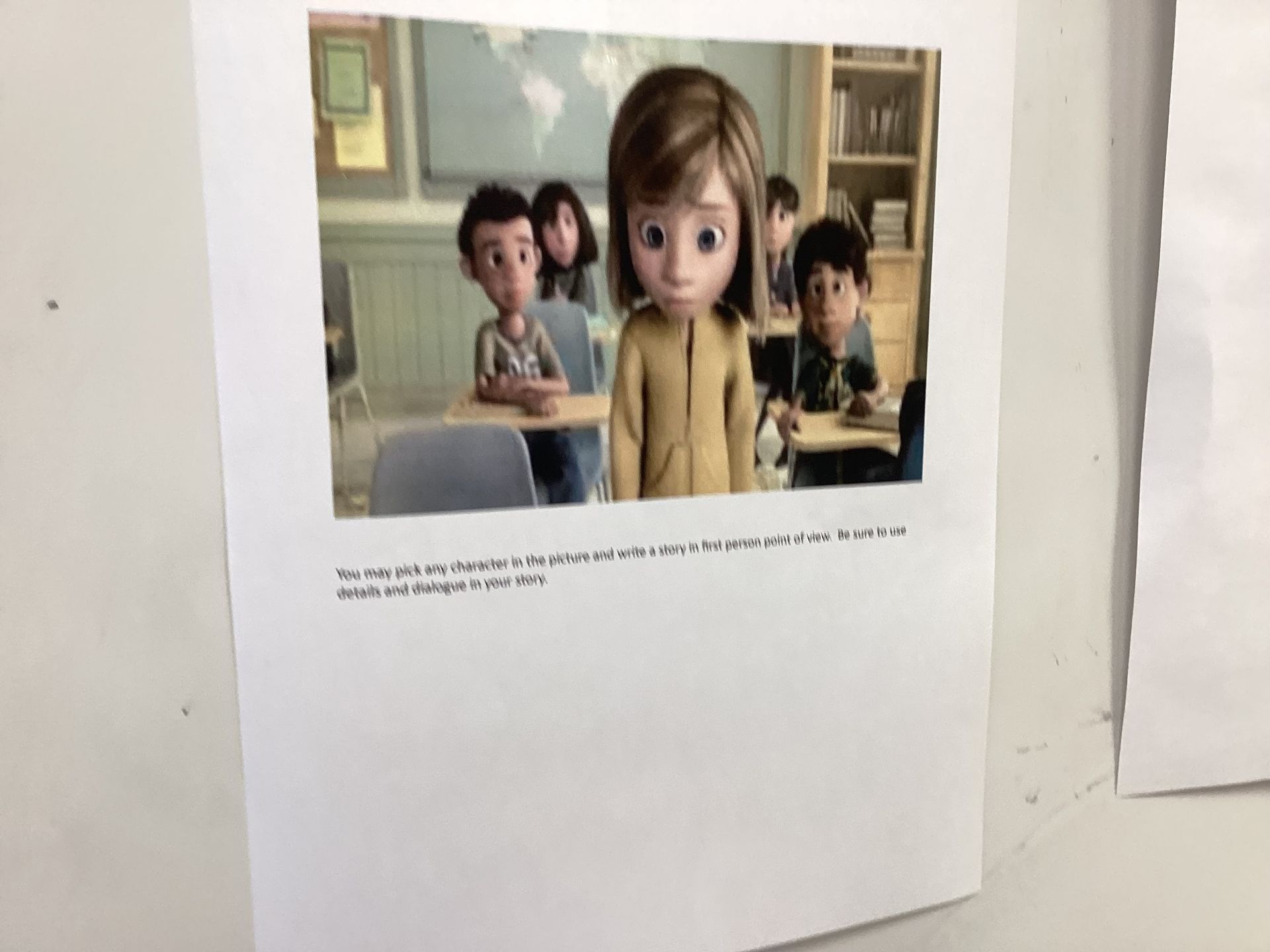 A printout of a scene from the movie Inside Out, showing several characters sitting at desks in a classroom. Sadness is in the front, looking forlorn. The text on the paper reads: "You may pick any character in the picture and write a story in first person point of view. Be sure to use details and dialogue in your story."