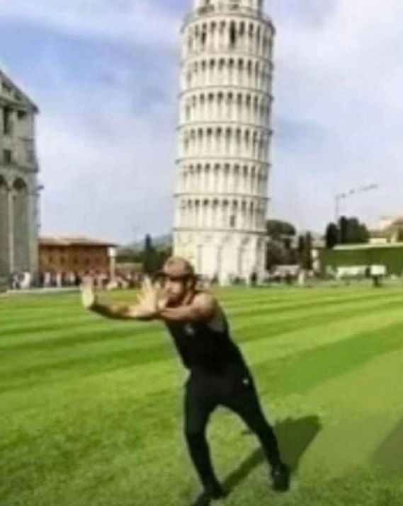 A man is positioned in front of the Leaning Tower of Pisa, with his arms extended as if he's trying to push it back to a straight position.